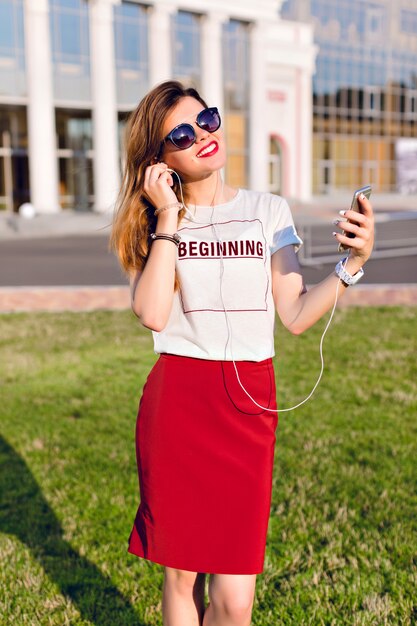 Retrato de una joven sonriente de pie sosteniendo un teléfono inteligente y escuchando música en auriculares