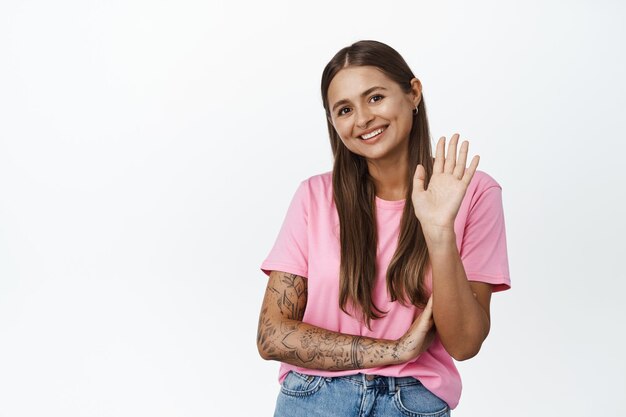 Retrato de una joven sonriente que agita su mano para saludar, saludando a la gente con expresión de cara feliz, de pie sobre fondo blanco.