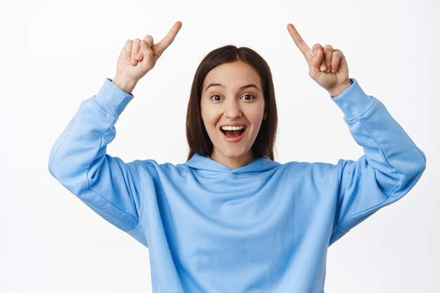 Retrato de una joven sonriente señalando con el dedo el logotipo de venta que muestra el anuncio de promoción de texto publicitario de pie en una sudadera con capucha contra el fondo blanco
