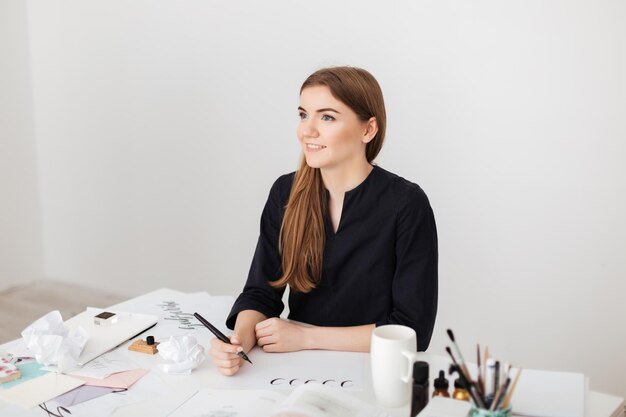 Retrato de una joven sonriente sentada en el escritorio blanco y escribiendo notas en papel mientras mira soñadoramente a un lado aislada