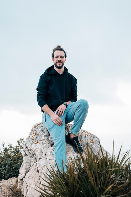 Retrato de un joven sonriente sentado en la roca mirando a la cámara contra el cielo azul