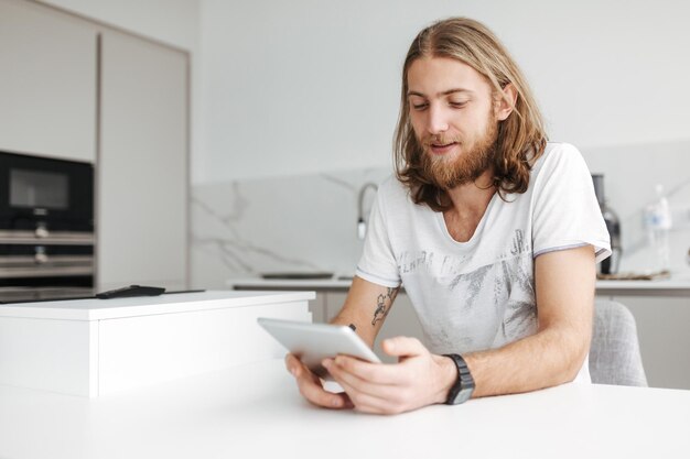 Retrato de un joven sonriente sentado y usando una tableta digital en la cocina de casa