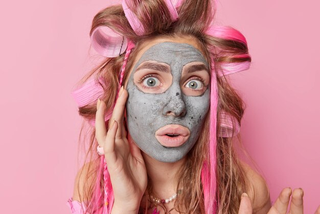 El retrato de una joven sorprendida aplica una máscara de arcilla hidratante en la cara para el tratamiento de la piel se somete a procedimientos antienvejecimiento usa rulos para el cabello se ve impresionado por la cámara aislada sobre una pared rosada