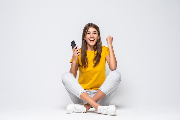 Retrato de una joven sorprendida que usa el teléfono mientras está sentado con las piernas cruzadas aislado en blanco