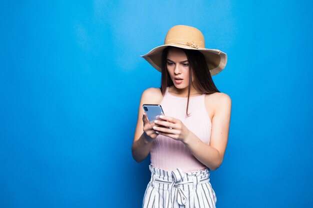 Retrato de una joven sorprendida con sombrero de verano mirando el teléfono móvil aislado sobre la pared azul.