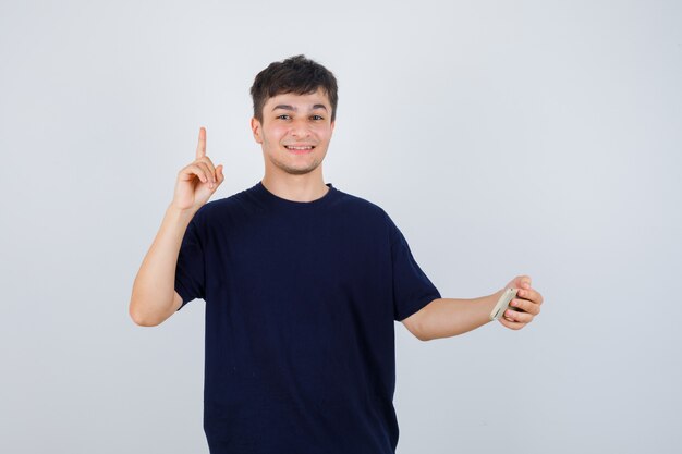 Retrato de joven sosteniendo un teléfono móvil, apuntando hacia arriba en una camiseta negra y mirando confiado vista frontal