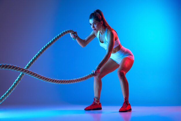 Retrato de joven spotive chica haciendo ejercicios con cuerda manteniendo el cuerpo en forma aislado sobre fondo azul en neón