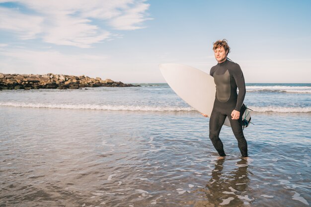 Retrato de joven surfista dejando el agua con tabla de surf bajo el brazo. Concepto de deporte y deporte acuático.