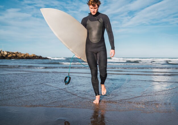 Retrato de joven surfista dejando el agua con tabla de surf bajo el brazo. Concepto de deporte y deporte acuático.