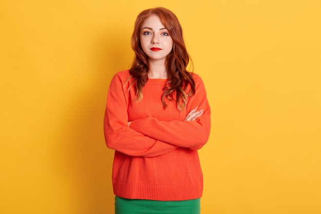 Retrato de joven tierna mujer europea de pelo rojo con mirada seria, vistiendo un suéter naranja, mirando a la cámara con expresión tranquila o triste