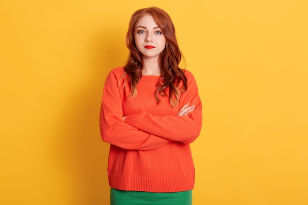 Retrato de joven tierna mujer europea de pelo rojo con mirada seria, vistiendo un suéter naranja, mirando a la cámara con expresión tranquila o triste