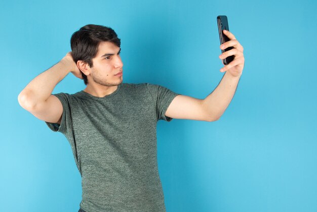 Retrato de un joven tomando selfie con teléfono móvil contra azul.