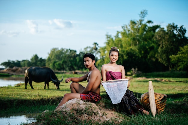 Retrato de joven en topless sentado cerca de una mujer bonita en ropa hermosa en el estilo de vida rural, trampa de pesca de bambú