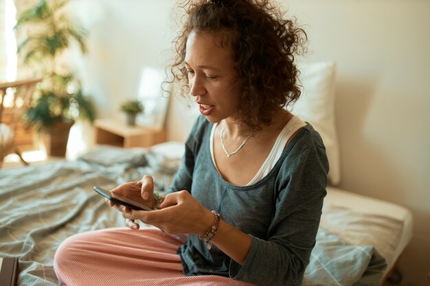 Retrato de joven trabajadora independiente latina vestida informalmente vendiendo productos en línea, sentada en la cama con el teléfono móvil, escribiendo mensajes de texto, charlando con el cliente