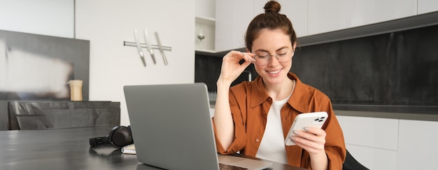 Retrato de una joven trabajadora independiente que responde a los clientes en su teléfono inteligente utilizando una computadora portátil para comprobar