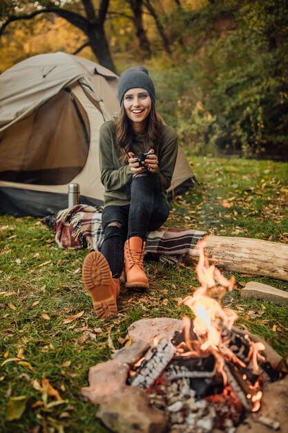 Retrato de joven turista hermosa sentada en el tronco en el bosque cerca de la tienda y el saco de dormir