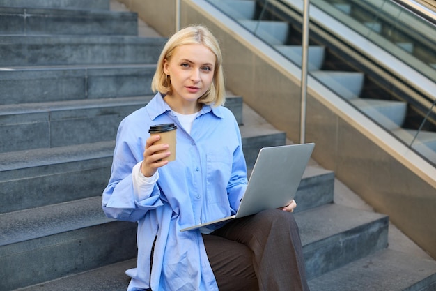 Retrato de una joven urbana sentada en las escaleras con una taza de café para llevar y trabajando en una computadora portátil