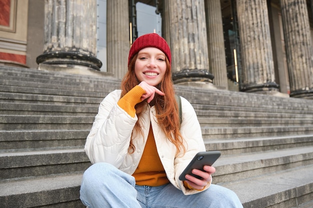 Retrato de una joven urbana con sombrero rojo sentada en las escaleras cerca del museo sostiene que el teléfono móvil se conecta a la pu