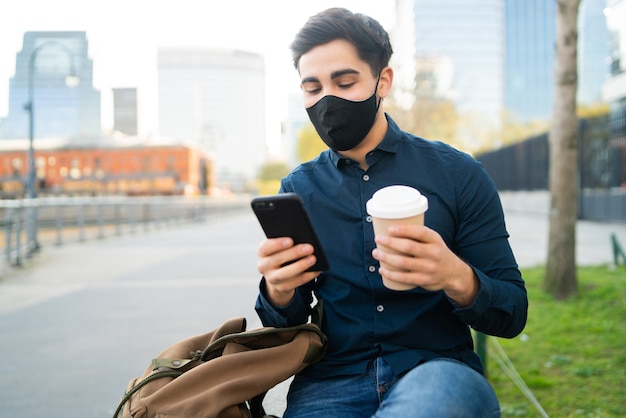 Retrato de joven usando su teléfono móvil y sosteniendo una taza de café mientras está sentado en un banco al aire libre