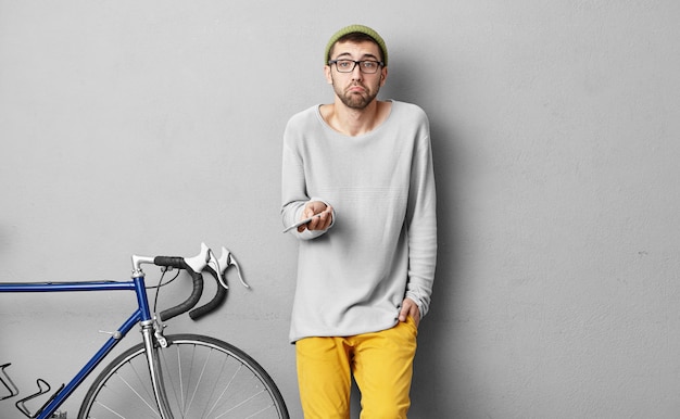 Retrato de un joven viajero masculino, con una carrera ciclista, sosteniendo un teléfono inteligente moderno en las manos, usando el navegador, tratando de encontrar la manera adecuada, teniendo una expresión dudosa mientras se pierde en un lugar desconocido