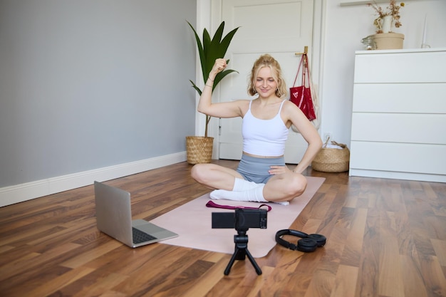 Foto gratuita retrato de una joven vlogger instructora de fitness que muestra ejercicios en la cámara grabándose a sí misma sentada