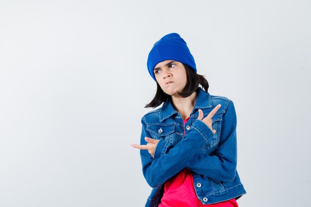 Retrato de jovencita apuntando a derecha e izquierda en chaqueta de mezclilla, gorro y mirando enfocado vista frontal