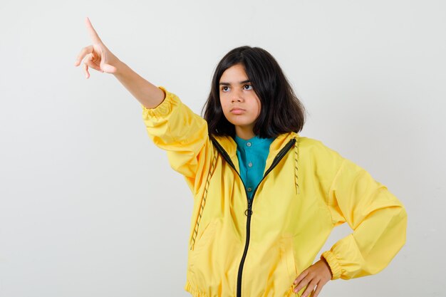 Retrato de jovencita apuntando hacia fuera en chaqueta amarilla y mirando sombría vista frontal