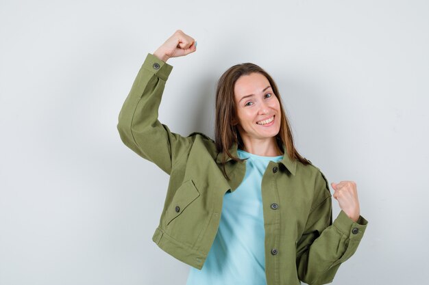 Retrato de jovencita mostrando gesto ganador en chaqueta verde y mirando feliz vista frontal