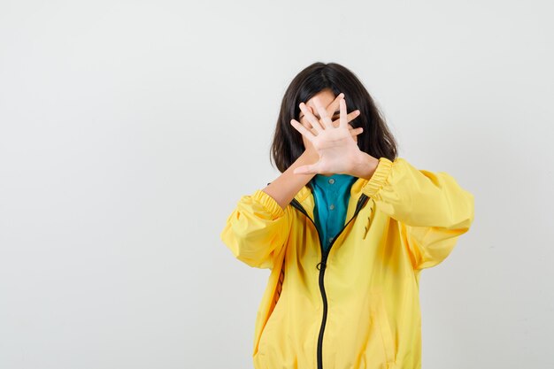 Retrato de jovencita mostrando gesto de parada, sosteniendo la mano en la cara con chaqueta amarilla y mirando asustado vista frontal