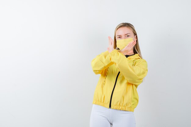 Retrato de jovencita mostrando gesto de rechazo en chaqueta, pantalón, máscara y mirando confiada vista frontal