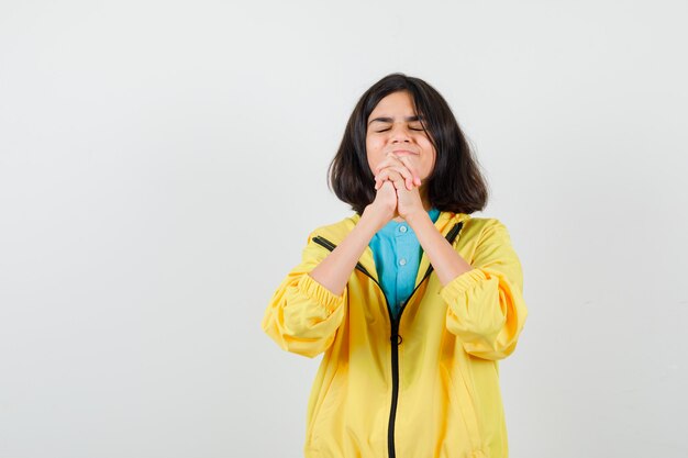 Retrato de jovencita mostrando las manos entrelazadas en gesto de súplica en chaqueta amarilla y mirando esperanzada vista frontal