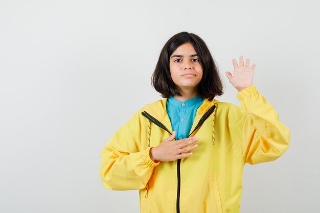 Retrato de jovencita mostrando la palma, sosteniendo la mano en el pecho con chaqueta amarilla y mirando confiada vista frontal