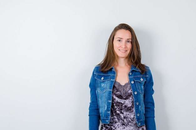 Retrato de jovencita posando mientras está de pie en blusa, chaqueta de mezclilla y mirando alegre vista frontal