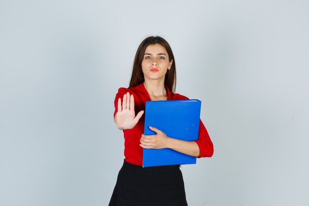 Retrato de jovencita sosteniendo la carpeta mientras muestra el gesto de parada en blusa roja