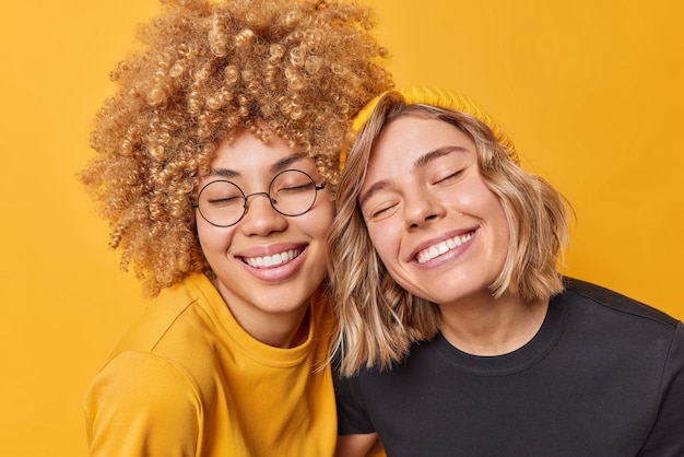 Retrato de jóvenes europeas felices que mantienen los ojos cerrados por la sonrisa de satisfacción que están de buen humor vestidas con camisetas informales expresan emociones positivas aisladas sobre el fondo amarillo