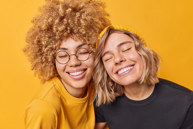 Retrato de jóvenes europeas felices que mantienen los ojos cerrados por la sonrisa de satisfacción que están de buen humor vestidas con camisetas informales expresan emociones positivas aisladas sobre el fondo amarillo