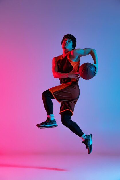 Retrato de jugador de baloncesto de niño activo en un entrenamiento de salto aislado sobre fondo rosa púrpura degradado