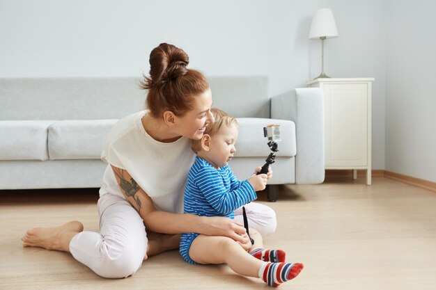 Retrato de lado de la feliz madre caucásica joven y su hijo sentados en el suelo en casa, haciendo selfie. Mujer sonriente en ropa blanca abrazando a su bebé. Niño tomando una foto con selfie stick.