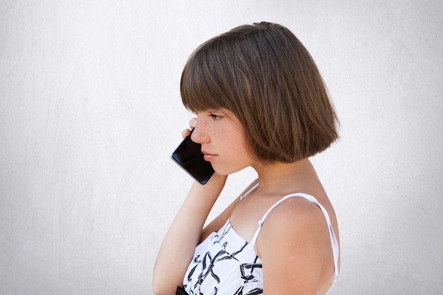 Retrato de lado de niña con cabello corto, vestido blanco, hablando por teléfono celular con expresión seria. Elegante niña posando en estudio con gadget moderno, aislado en blanco