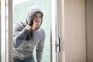 Foto gratuita retrato de un ladrón con capucha y guantes mirando dentro de una casa a través de una puerta de cristal