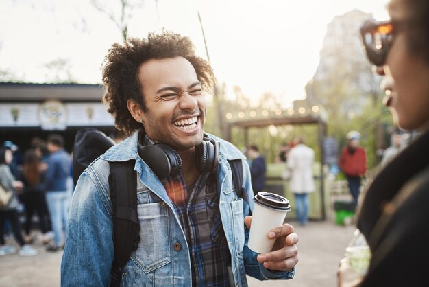 Retrato lateral de un chico afroamericano lindo y de moda con peinado afro que se ríe a carcajadas de una broma mientras habla con un amigo en el parque tomando café y siendo optimista Concepto de amistad