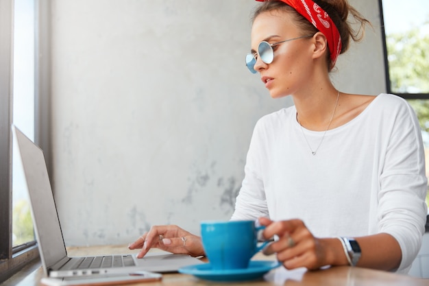 Foto gratuita el retrato lateral de una joven y concentrada diseñadora gráfica bastante femenina viste tonos de moda, trabaja en una computadora portátil moderna y toma café, pasa el descanso para cenar en la cafetería, prepara el trabajo del proyecto.