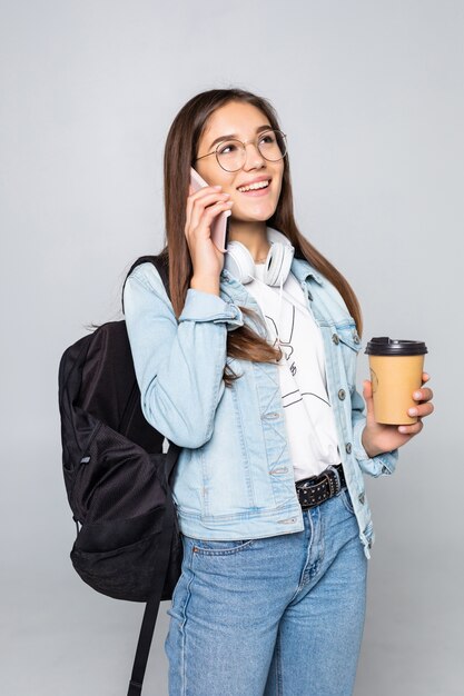 Retrato lateral de mujer joven estudiante hablar con teléfono inteligente, sosteniendo café para llevar taza aislado en la pared gris