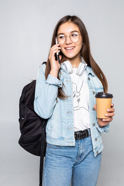 Retrato lateral de mujer joven estudiante hablar con teléfono inteligente, sosteniendo café para llevar taza aislado en la pared gris