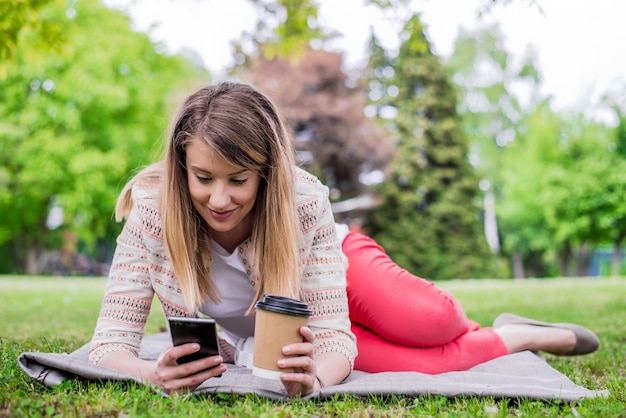 Retrato lateral de la mujer riendo que miente en hierba afuera con el teléfono móvil. Chica usando un teléfono inteligente en la hierba de un parque con un fondo verde