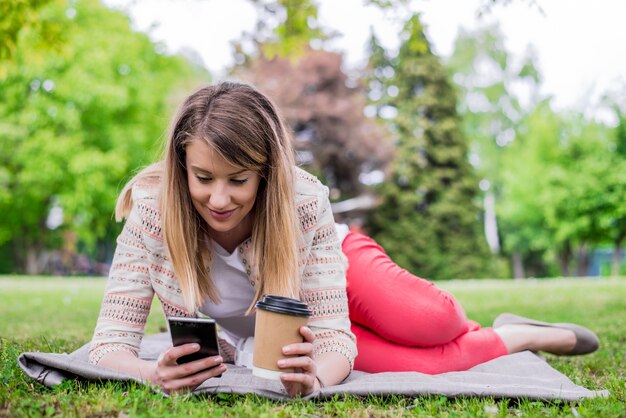 Retrato lateral de la mujer riendo que miente en hierba afuera con el teléfono móvil. Chica usando un teléfono inteligente en la hierba de un parque con un fondo verde