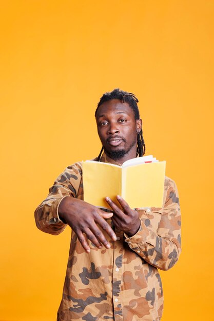 Retrato de un lector serio sosteniendo un libro de literatura, leyendo una historia de ficción en un estudio sobre fondo amarillo. Hombre afroamericano concentrado aprendiendo antes de comenzar a trabajar en la tarea universitaria