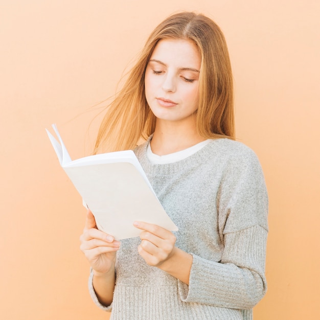 Retrato de un libro de lectura rubio de la mujer joven contra el contexto del color del melocotón