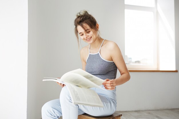 Retrato del libro de lectura sonriente de la mujer feliz hermosa joven que se sienta en silla sobre la pared blanca en casa.