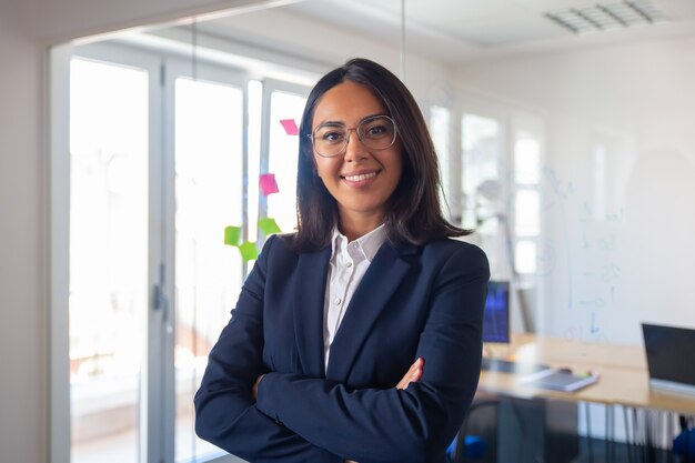 Retrato de líder empresarial latino confiado. Joven empresaria en traje y gafas posando con los brazos cruzados, mirando a cámara y sonriendo. Concepto de liderazgo femenino